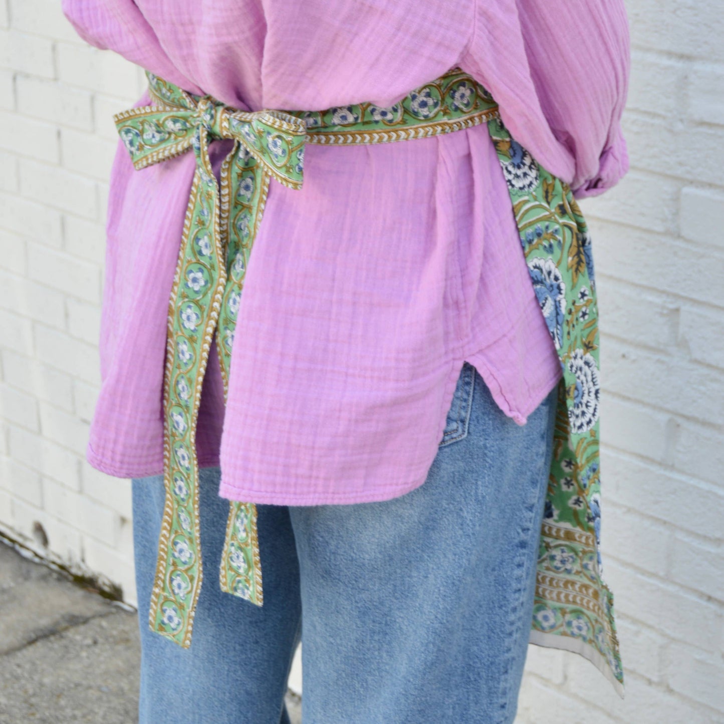 Person wearing a pink top with a patterned hand blocked green and blue apron against a white brick wall.