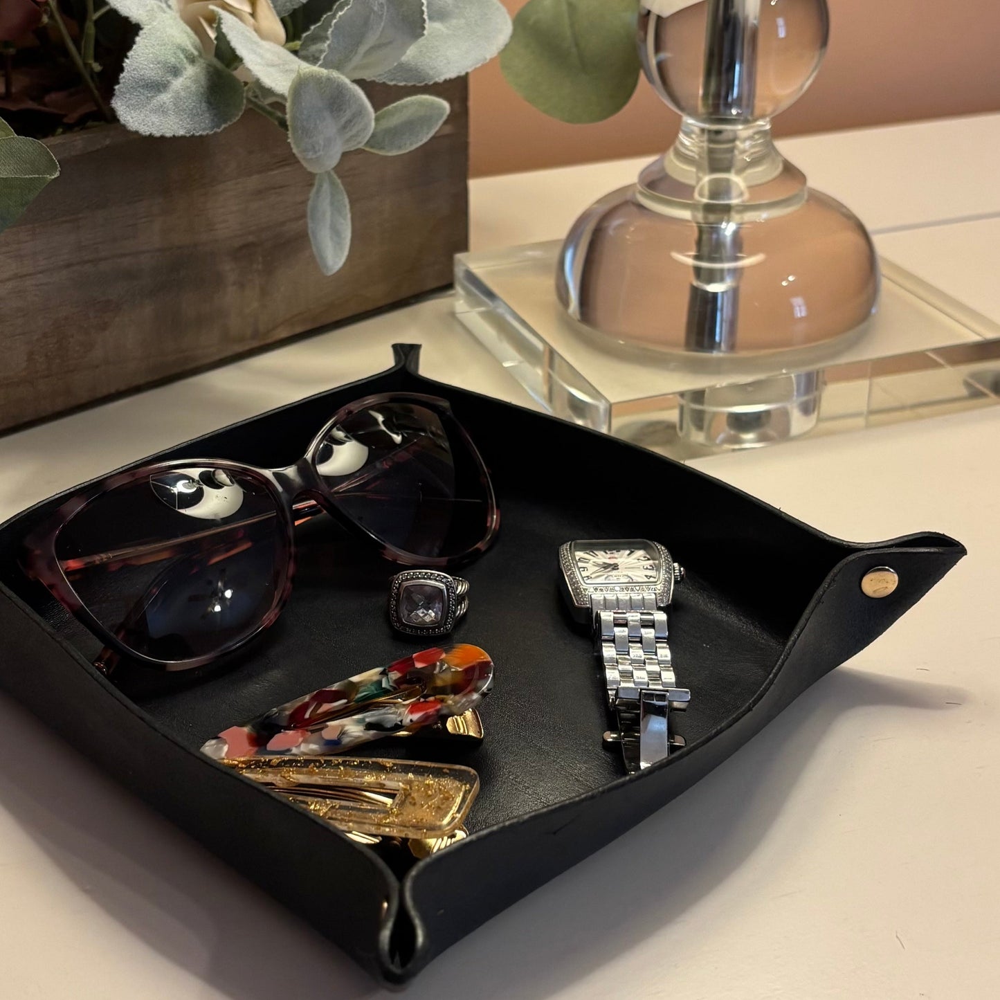 Navy catchall tray with sunglasses, watch, and ring on a surface with a decorative lamp and plant in the background.
