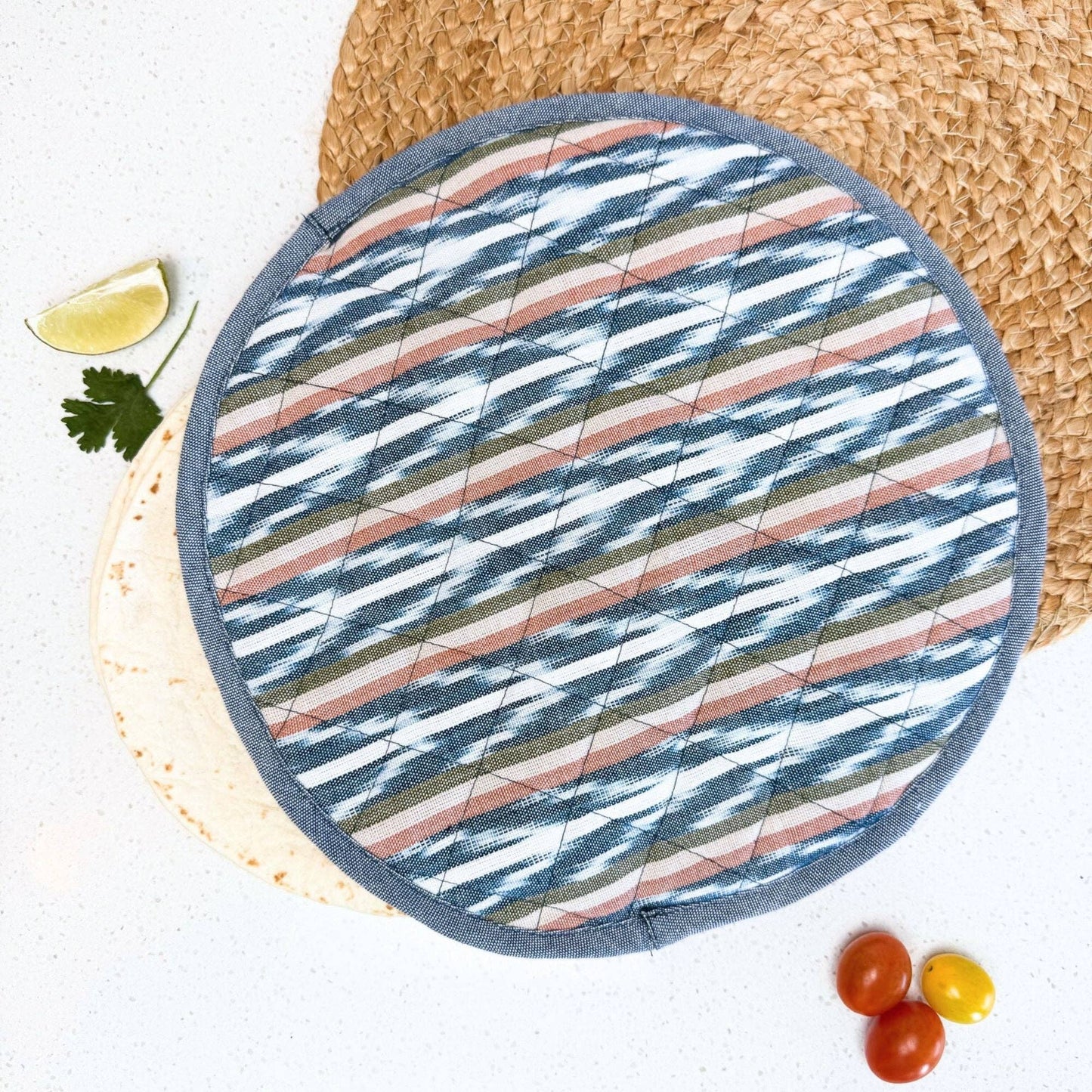 Round quilted tortilla warmer with striped pattern on a white kitchen surface styled with a straw placemat, a line wedge and cherry tomatoes.