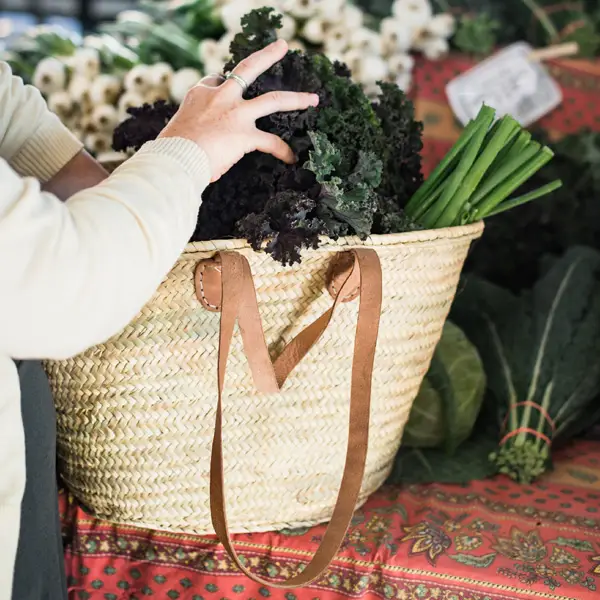 the arms and hands of a women placing kale into a straw french market basket