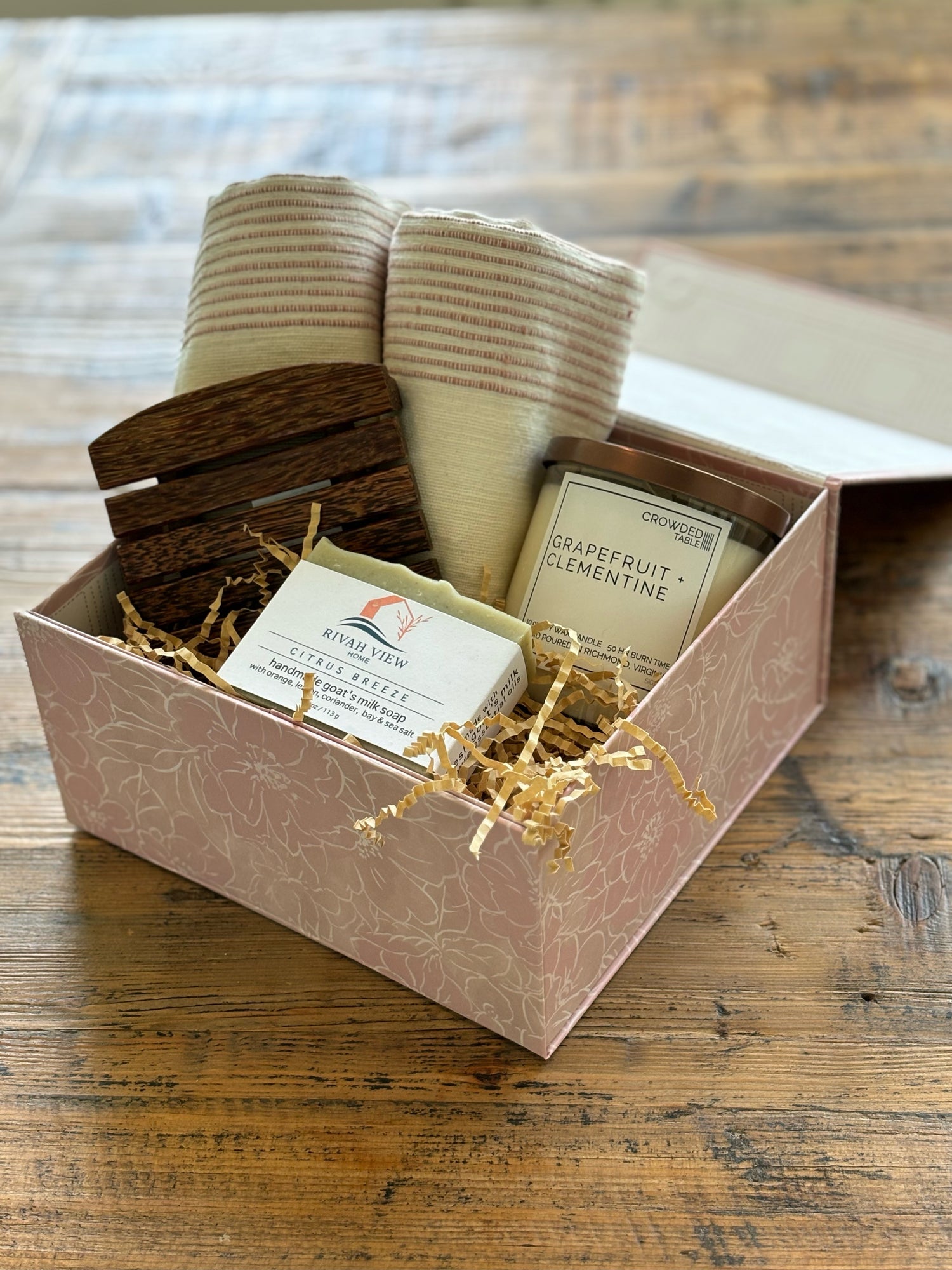 pink gift box filled with handmade wood soap dish, two rolled up stripped hand towels, a candle and a goat milk soap bar