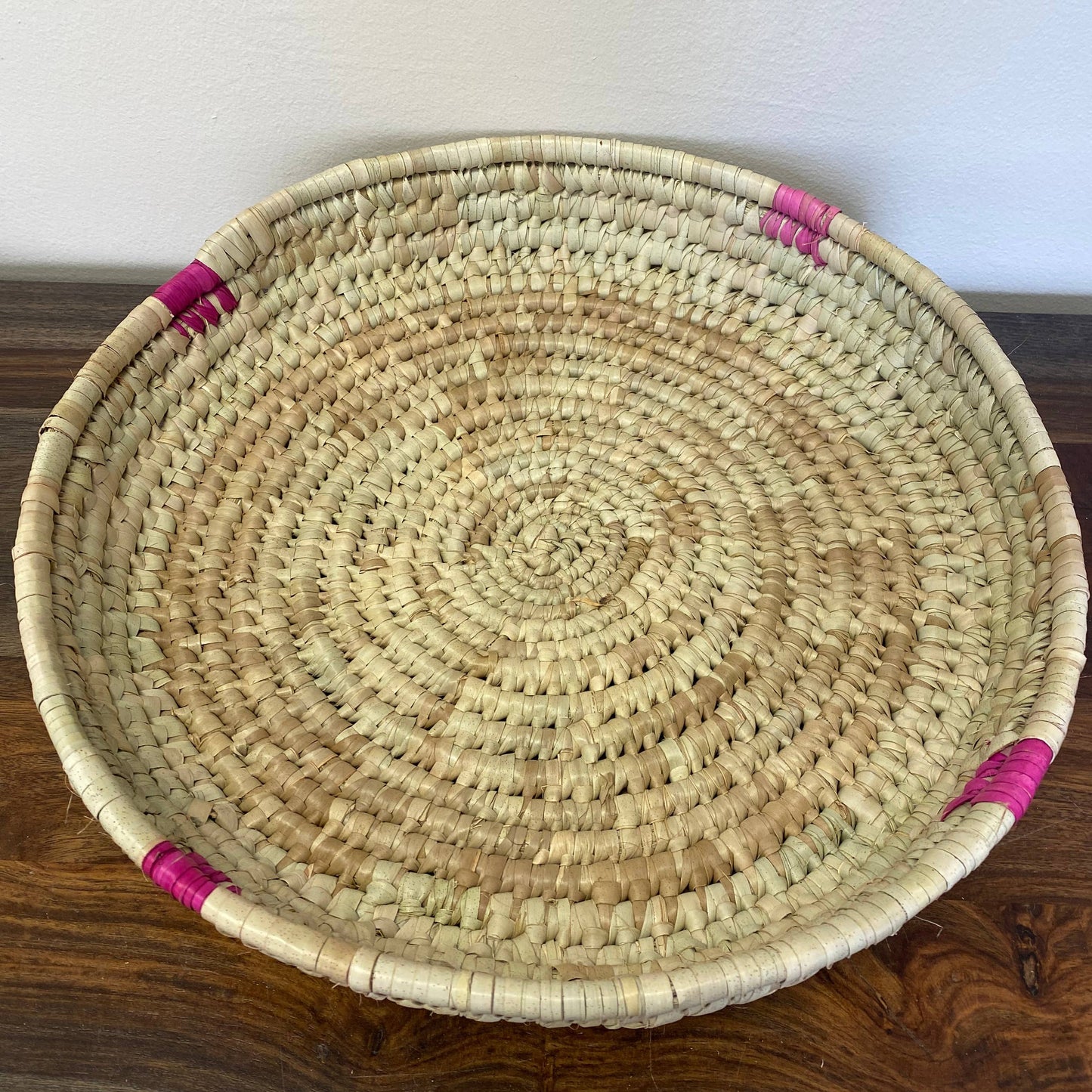 A round, beige palm leaf tray with pink detailing, placed on a wooden surface against a white wall.