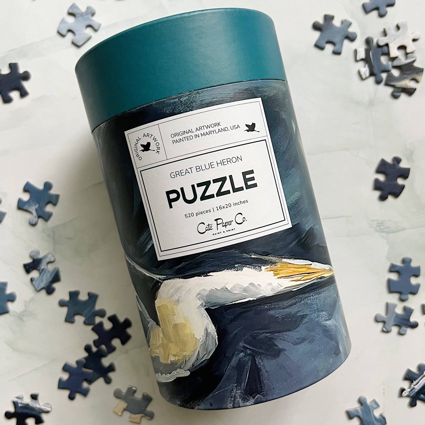 Blue Heron Jigsaw puzzle packaging with a label on a white background with scattered puzzle pieces.