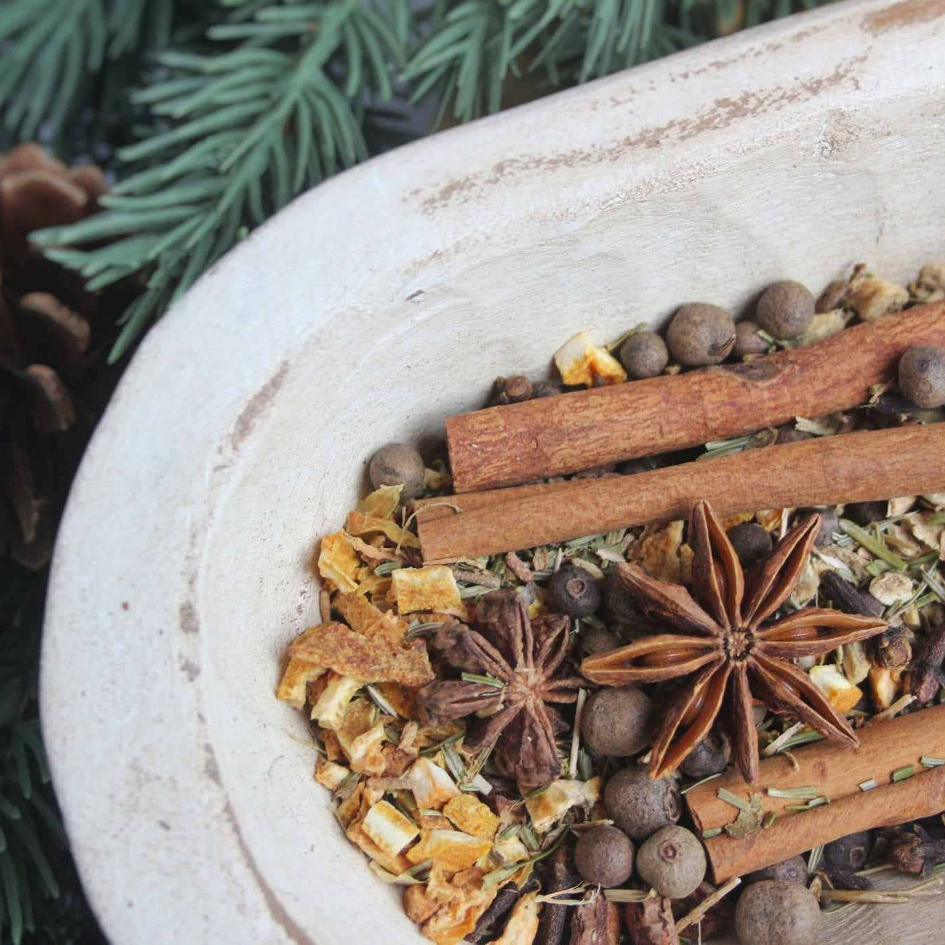 Potpourri spices including cinnamon sticks, star anise, and cloves in a bowl with pine cones and branches.