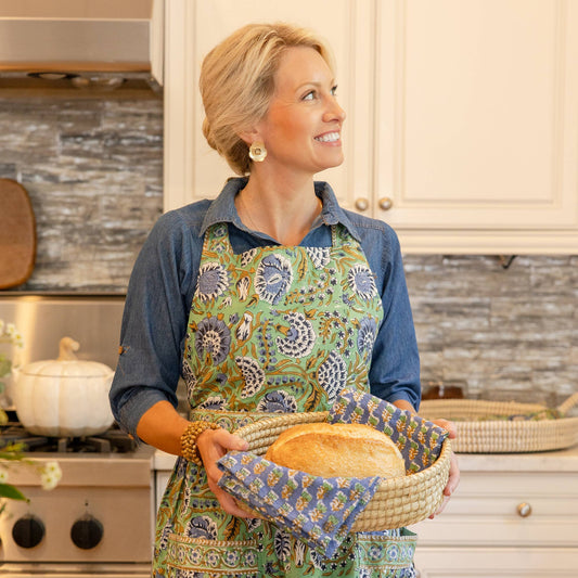 Woman in a kitchen holding a basket of bread wearing a patterned blue and mint hand blocked printed apron.