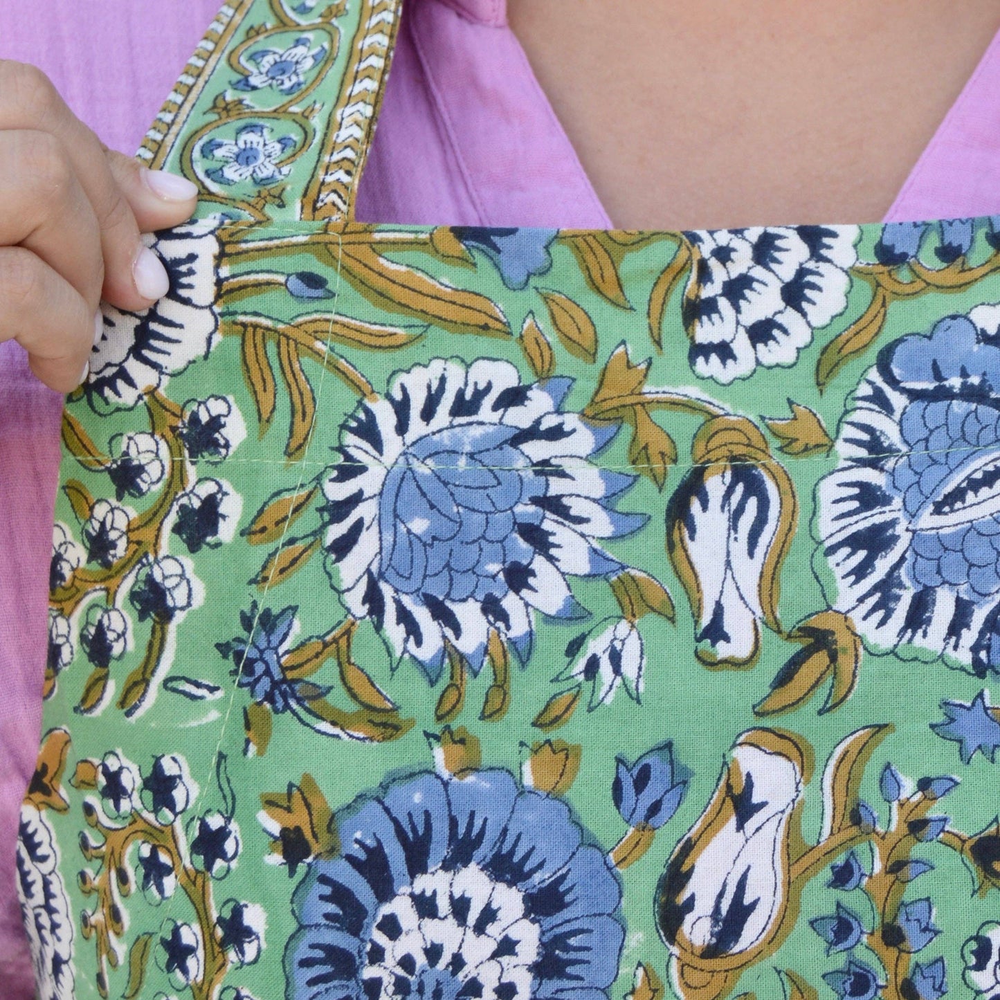 Green floral apron with blue and white hand blocked flowers held by a person wearing a pink shirt.