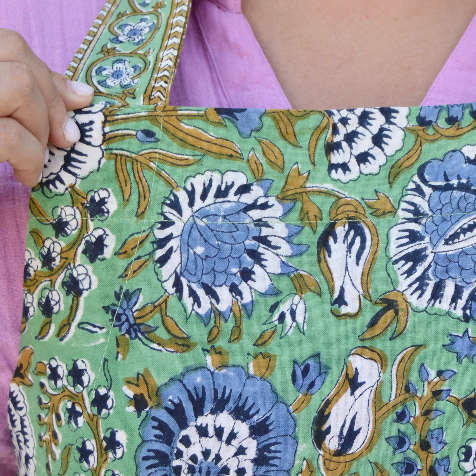 Green floral apron with blue and white hand blocked flowers held by a person wearing a pink shirt.