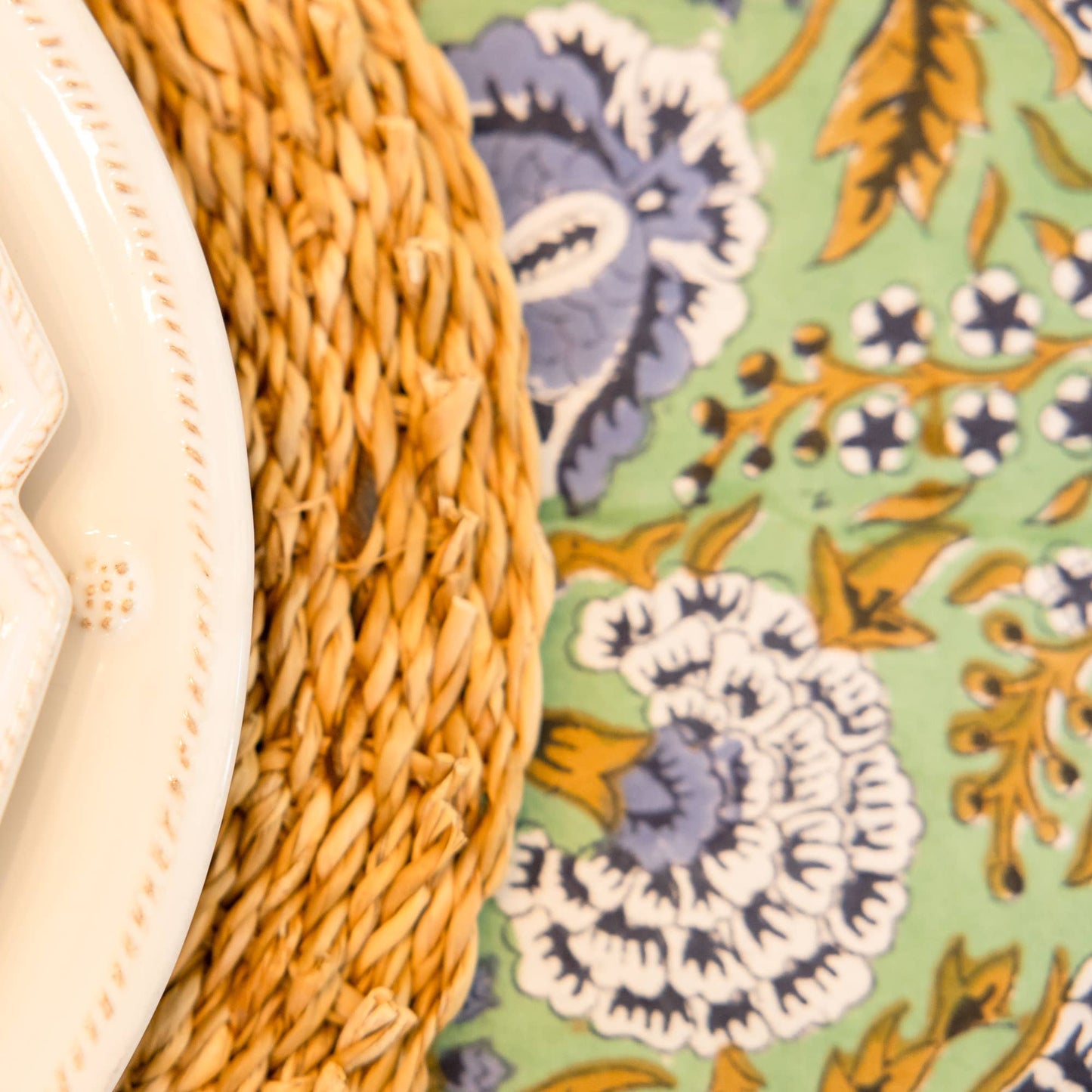 Close-up of a textured white plate, woven charger, and floral-patterned mint colored hand blocked tablecloth.