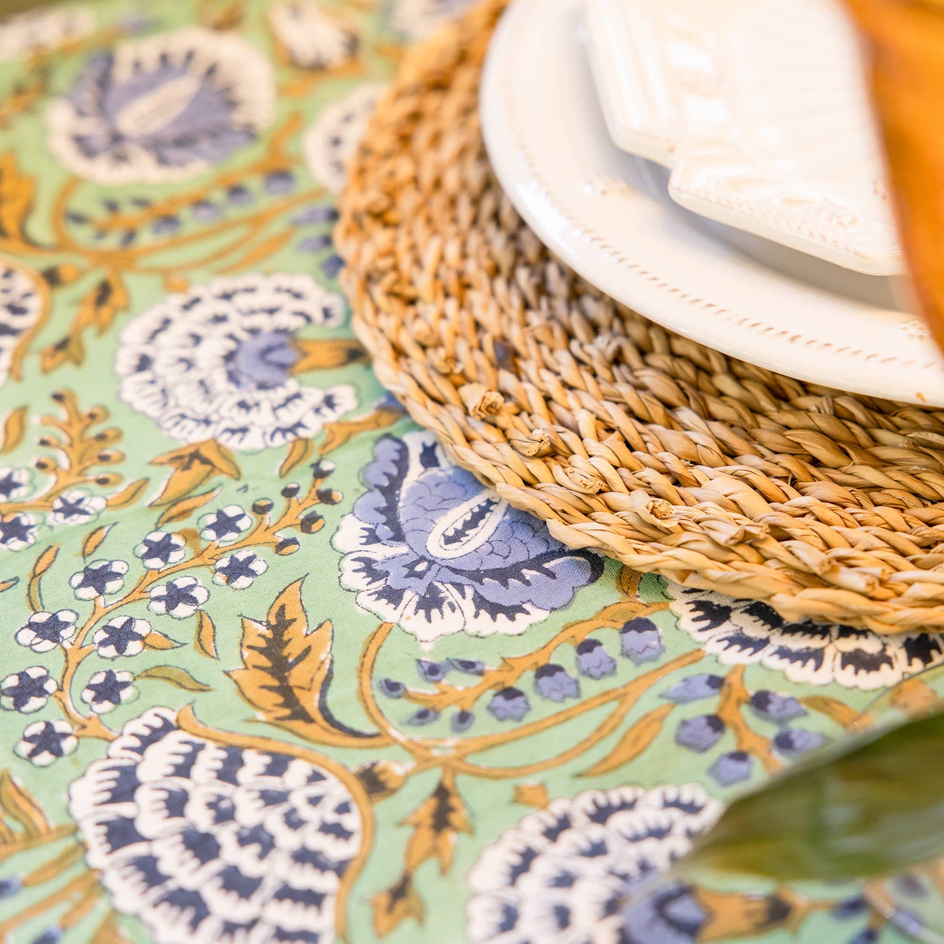 Close-up of a table setting with a floral hand blocked tablecloth, woven placemat, and glass.
