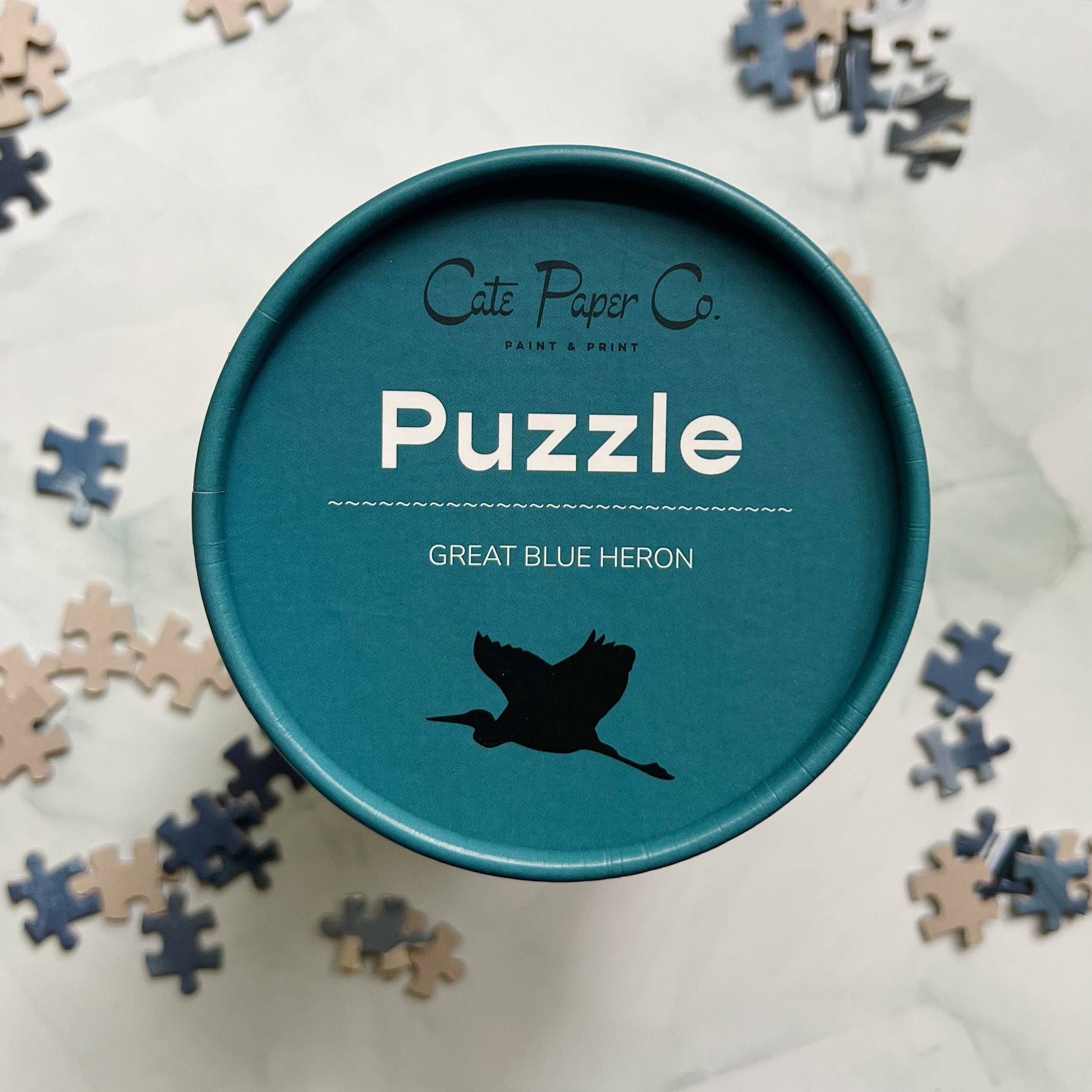 Blue Heron puzzle box with 'Cate & Paper Co' branding and scattered puzzle pieces on a light background