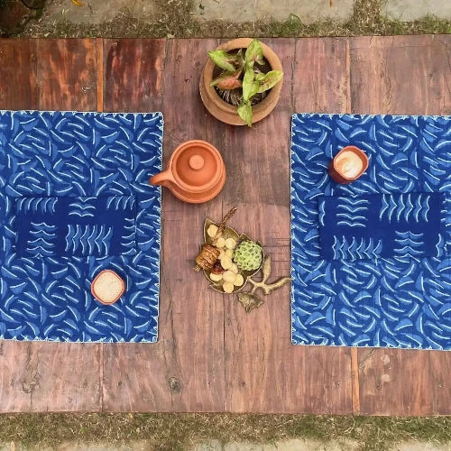 Two blue patterned placemats on a wooden table with a small plant and ceramic items.