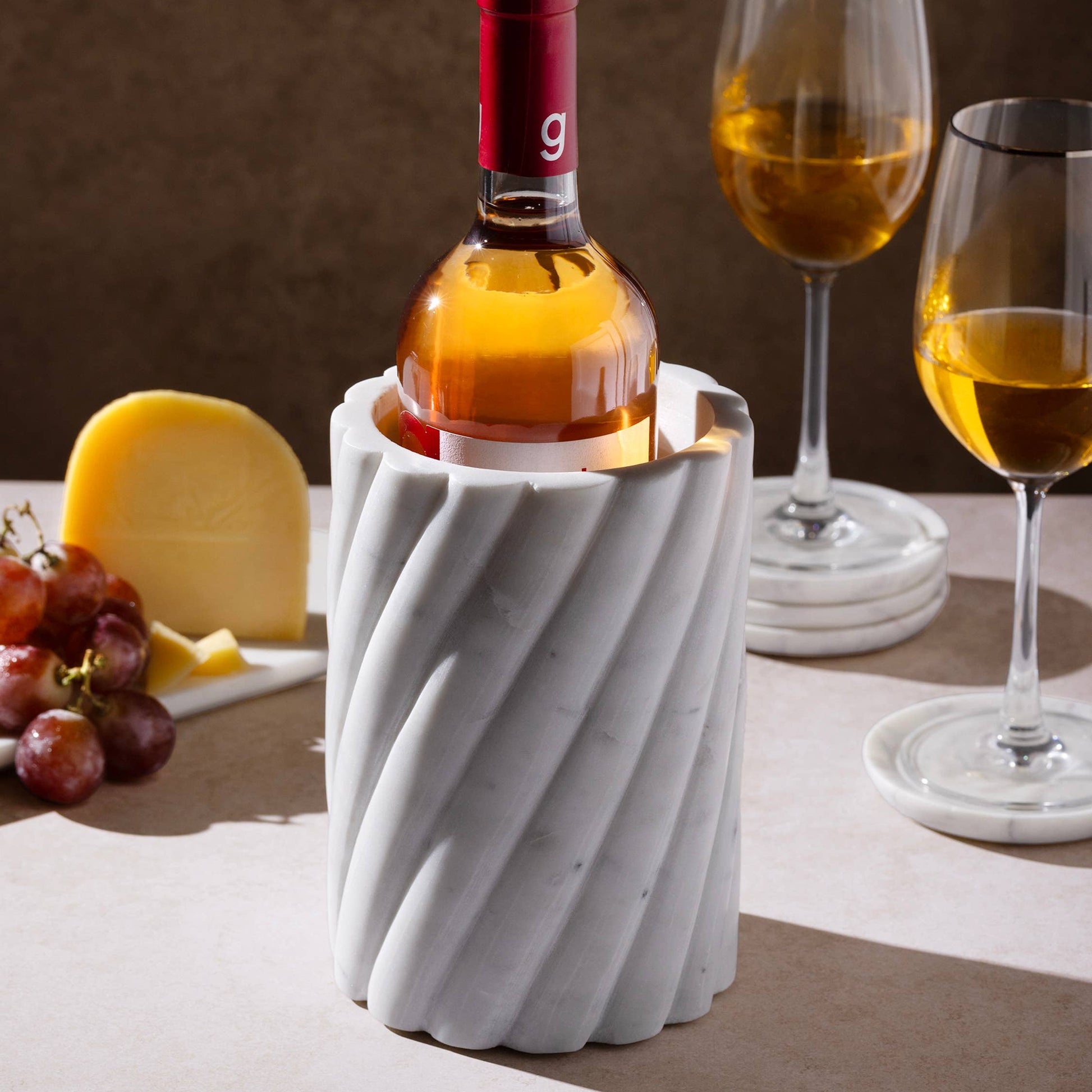 Bottle of wine in a decorative white marble holder with two glasses and cheese on a table.