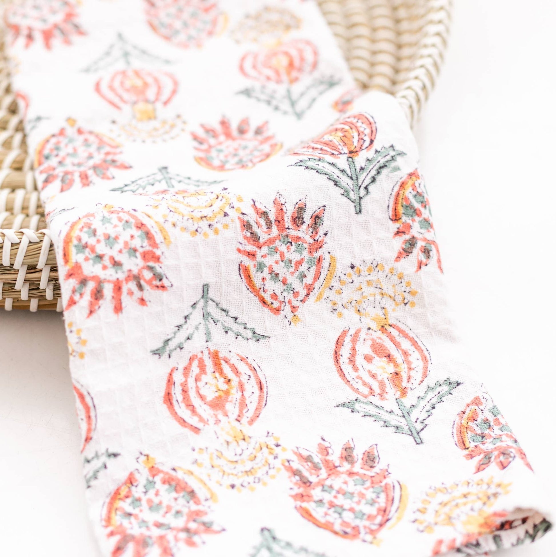 A folded beige cotton waffle tea towel with a multicolored floral block print design, displayed in front of a woven basket.