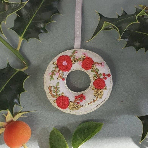 A circular embroidered floral ornament with red roses and green leaves on a white base, hanging from a light pink ribbon, styled with holly leaves and a rose on a gray background.