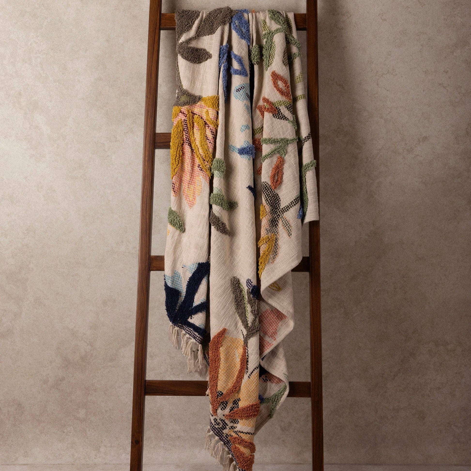 Handcrafted decorative blanket with floral patterns draped over a wooden ladder against a neutral wall.